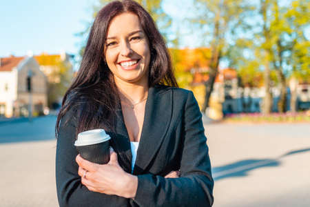 Happy 30s beautiful caucasian business woman drinking hot coffee holding paper cup while walking in city sunset park.の写真素材