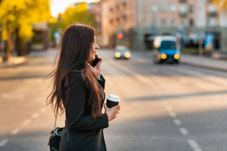 Young business woman talking on the phone holding coffee paper cup,crossing street blurred background. Fashion business photo of beautiful girl in black suite with phone and cup of coffee.Side view.の写真素材