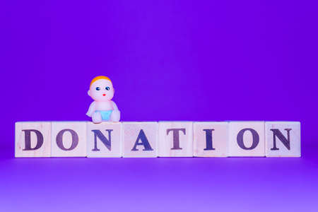 Business and finance. Wooden cubes with the inscription DONATION.Miniature toy baby is sitting on the cube.Purple,veri peri color indoors background.Copy space.の写真素材