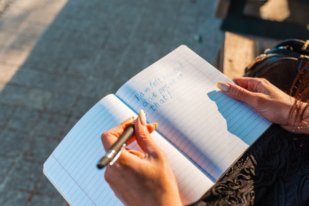 Woman writes in a notebook, holding pen in her left hand.Outdoors summer evening park. Left-handers Day August 13th.Closeup.の写真素材