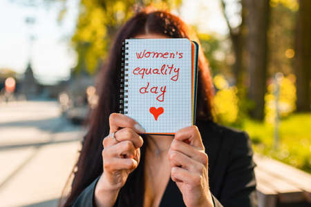 Young caucasian woman outdoors showing a notebook paper in front of her face with the text womens equality written in it.Summer sunset park evening.Closeup.Selective focus.の写真素材