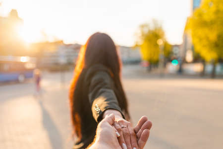 Happy brunette girl turn away face holding boyfriend's hand on a street at sunset on a warm spring,summer evening.Follow-me concept.Selective focus.の写真素材