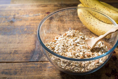 Cooking granola at morning: bowl with rolled oats, chopped nuts and spices, wooden spoon and fresh bananas on rustic wooden table. Copy spaceの写真素材