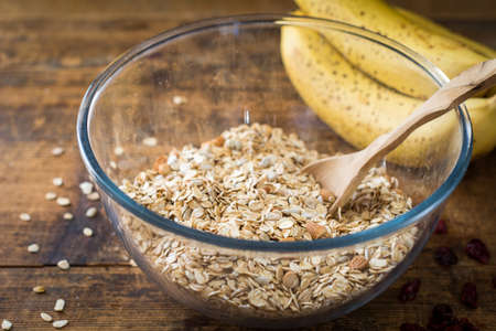 Cooking granola at morning: bowl with rolled oats, chopped nuts and spices, wooden spoon and fresh bananas on rustic wooden tableの写真素材