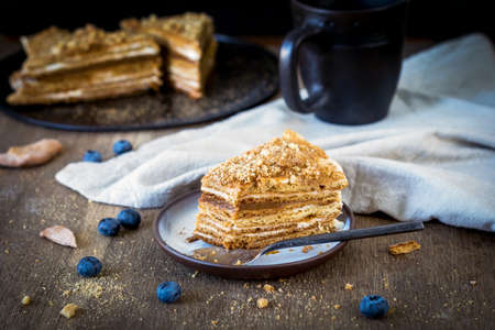 Slice of homemade medovik layered honey cake, traditional russian cake with cream and caramel on plate on rustic wooden table. Horizontal composition, selective focus, warm toneの写真素材