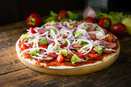 Thin crust pizza with sausages, pepper, tomatoes, broccoli and cheese on round wooden boardの写真素材