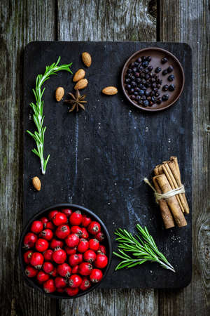 Food ingredients for cooking on chopping board with copy space. Fresh rosemary, cinnamon sticks, allspice, almonds, star anise and red berries on black textured background. Winter Christmas foodの写真素材