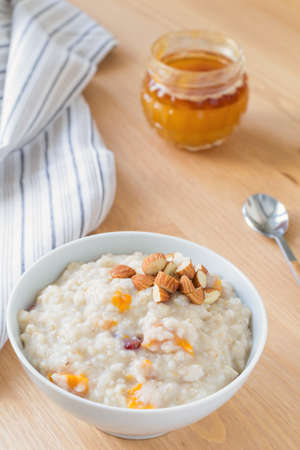 Oatmeal porridge with dried apricots and chopped almonds on bright wooden table. Jar of honey on background. Selective focus. Bright healthy breakfast imageの写真素材