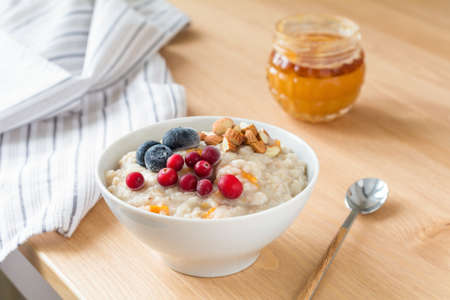 Oatmeal porridge with dried apricots, blueberries, cranberries and chopped almonds on bright wooden table. Jar of honey on background. Selective focus. Bright healthy breakfast imageの写真素材