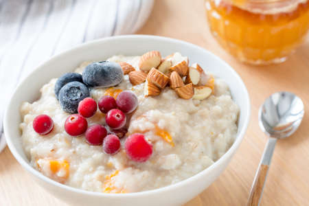 Oatmeal porridge with dried apricots, blueberries, cranberries and chopped almonds on bright wooden table. Jar of honey on background. Selective focus. Bright healthy breakfast imageの写真素材