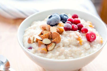 Oatmeal porridge with dried apricots, blueberries, cranberries and chopped almonds on bright wooden table. Jar of honey on background. Selective focus. Bright healthy breakfast imageの写真素材