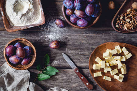 Ingredients for plum pie: fresh plums, walnuts, flour with sugar and chopped butter on rustic wooden table. Overhead viewの写真素材
