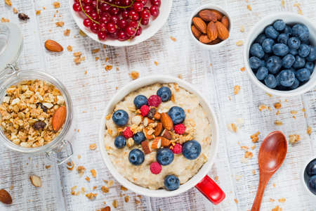 Bowl of oatmeal porridge with fresh blueberries, raspberries, muesli and almonds on wooden table. Jar of granola, red currants, fresh blueberries and almonds on side. Healthy breakfast conceptの写真素材