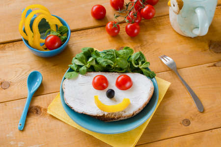 Breakfast meal for kid: funny smiley face sandwich with vegetables, soft white cheese and salad on blue plate.の写真素材
