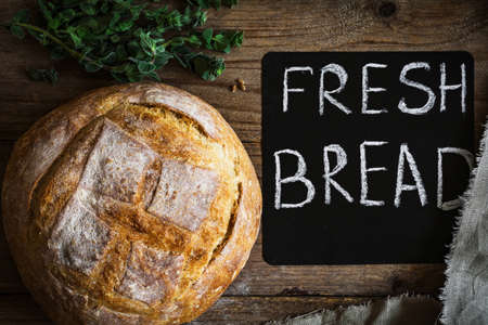 Round loaf of bread, freshly baked sourdough bread on wooden background. Chalk board and bunch of fresh oregano springs on sideの写真素材
