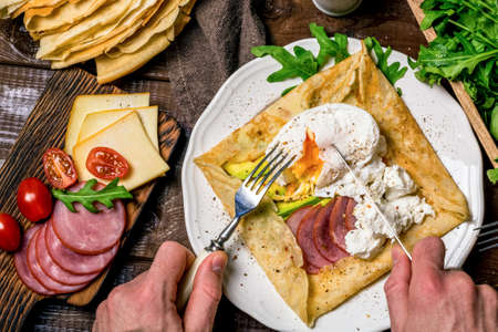 Man eating crepe galette with meat, avocado, soft white cheese and poached egg on white plate. Sliced ??yellow cheese, pastrami, cherry tomatoes, green salad and stack of crepes on side. Top viewの写真素材