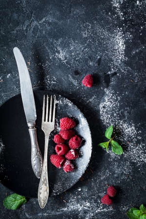 Black plate, vintage cutlery, fresh raspberries and mint leaf over dark stone background with copy space for text. Top view, vertical imageの写真素材