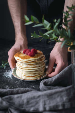 Stack of pancakes. Man hands serving pancakes with raspberries. Beautiful food still life. Natural light, slightly toned imageの写真素材