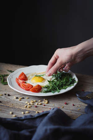 Breakfast with fried egg, green salad and fresh tomatoes. Female hand decorating plate with breakfast. Selective focus, rustic styleの写真素材
