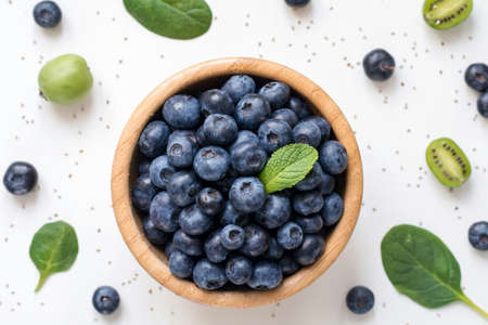 Blueberries in wooden bowl, top view. Flat lay or food pattern with blueberries, baby spinach, baby kiwi and chia seeds on white backgroundの写真素材