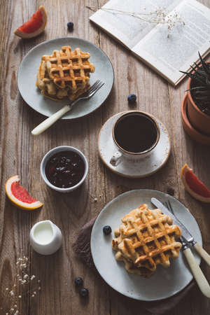 Waffles with jam and fruits, cup of coffee, cream and open book on rustic wooden table. Top view. Breakfast, morning conceptの写真素材