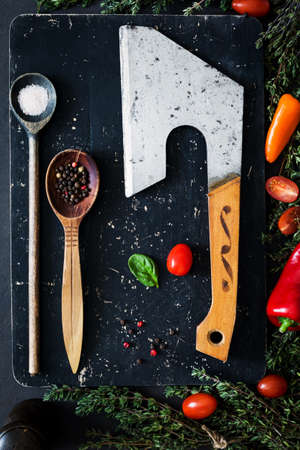 Old meat cleaver, spices, wooden spoon with spices, fresh greens and vegetables for cooking on black textured wooden cutting board. Top viewの写真素材
