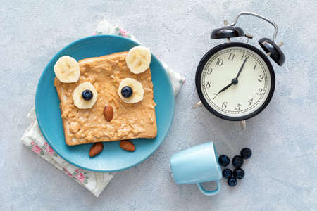 Funny owl toast with peanut butter, banana, blueberries and almonds on a blue plate, a clock and tiny cup of blueberries. Top view of healthy cute and healthy breakfast for kids. Good morning conceptの写真素材