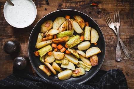 Roasted vegetables, potato wedges, carrots, onion, garlic and zucchini in pan with tzatziki sauce on rustic wooden table.の写真素材