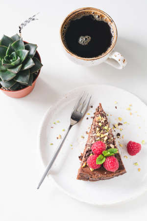 Slice of chocolate cake with raspberries and nuts, cup of black coffee and succulent plant stone rose on white table. Feminine breakfast food photographyの写真素材