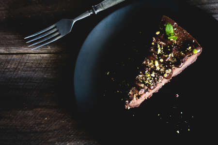Dark chocolate cheesecake decorated with chopped pistachios and mint leaf on black plate on wooden table. Top view, toned imageの写真素材
