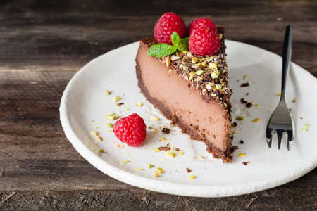 Slice of chocolate cheesecake decorated with pistachios, raspberries and mint leaf on white plate on wooden background.の写真素材