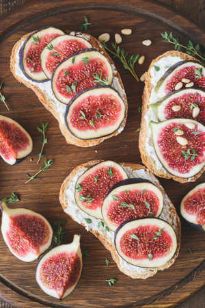 Bruschetta with fresh purple figs, ricotta cheese and honey on wooden cutting board garnished with fresh thyme and pine nuts. Healthy gourmet snack or appetizer. Table top viewの写真素材