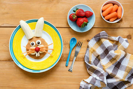 Easter breakfast for kids. Easter bunny shaped pancake with fruits on yellow plate. Colorful meal for children on wooden table, top viewの写真素材