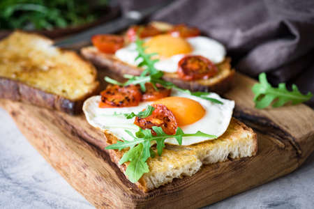 Healthy breakfast toast with egg, roasted tomato and arugula on rustic wooden cutting board. Closeup viewの写真素材