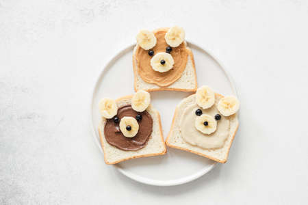Funny animal face toasts with nut butter, banana and blueberries on white plate. Kids meal. Vegan,...
