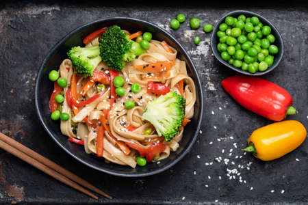 Asian stir fry with udon noodles and vegetables. Broccoli, green pea, pepper, carrots and pasta in black bowl. Top viewの写真素材