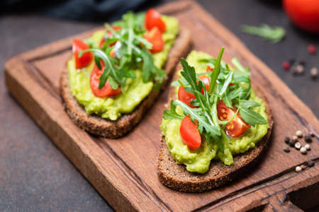 Healthy vegan toast with avocado, tomato, arugula on wooden serving board, closeup view, horizontal image. Snack, lunch or vegan breakfastの写真素材