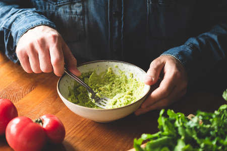 Man mashing avocado in bowl, wooden table background. Food preparation image, healthy vegan and vegetarian foodの写真素材