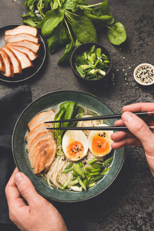 Eating japanese ramen noodle soup with chicken. Hand holding chopsticks above bowl of asian soup. Black concrete backgroundの写真素材