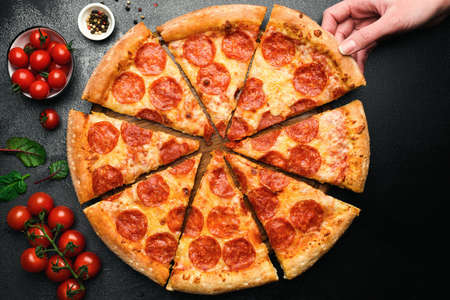 Hand picks slice of delicious pepperoni pizza. Table top view. On black concrete background. Concept...