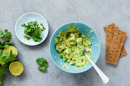 Mashed avocado in bowl and rye crispbreads. Making guacamole sauce, healthy vegetarian appetizer or...