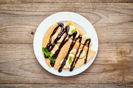 Crepes or blini with banana slices and chocolate sauce on white plate, wooden planksの写真素材