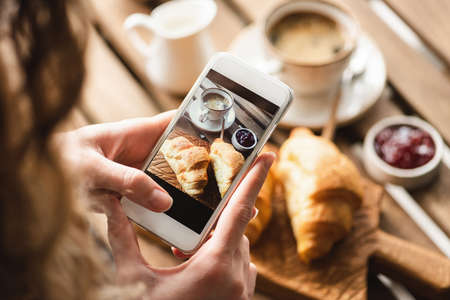 Woman taking picture of breakfast in cafe. Croissant, coffee with cream and jam on wooden table.の写真素材
