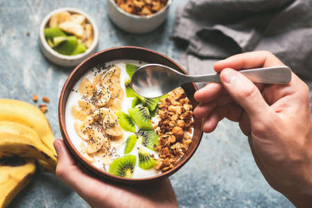 Eating healthy breakfast yogurt bowl with granola and fruits kiwi, banana, coconut.