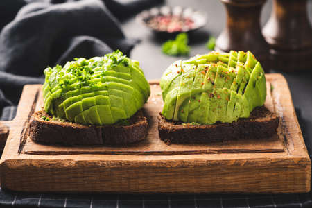 Rye bread toast with avocado garnished with spices and chopped parsley on a wooden cutting board....