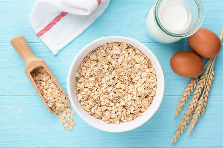Oat flakes or rolled oats in bowl, bottle of milk and chicken eggs. Blue wooden table background....