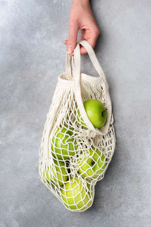 Hand holding mesh bag with green organic apples over grey concreteの写真素材
