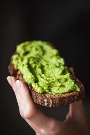 Hand holding rye bread toast with mashed avocado over black background. Healthy eating, vegan...