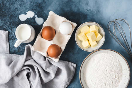 Ingredients for baking on blue background. Eggs, cubed butter, milk flour and whisker. Top view