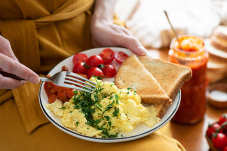 Eating scrambled eggs with baked beans and bread toast. Female hands holding plate with breakfast...